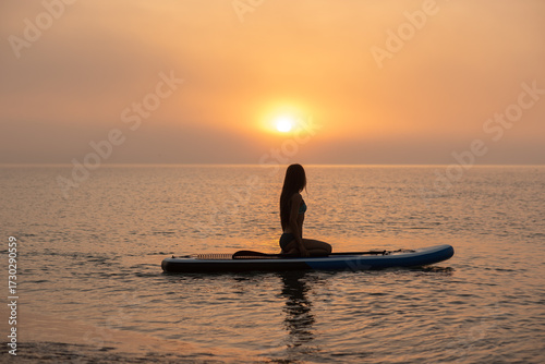 Fototapeta Naklejka Na Ścianę i Meble -  A beautiful, slender, athletic girl is relaxing on a SUP in the open sea at dawn on a sunny summer day.