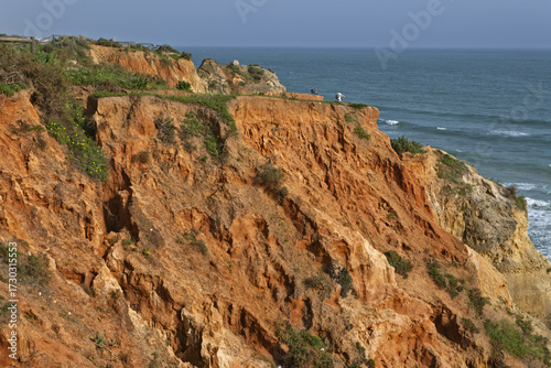 coastal view with golden cliffs, sandy beach, and turquoise ocean waves. The cliffs feature layered rock and sparse vegetation, with buildings visible on top in Portimao, Algarve, Portugal