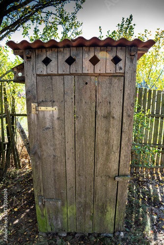 Vertical image, simple wooden outhouse near a broken wooden fence.