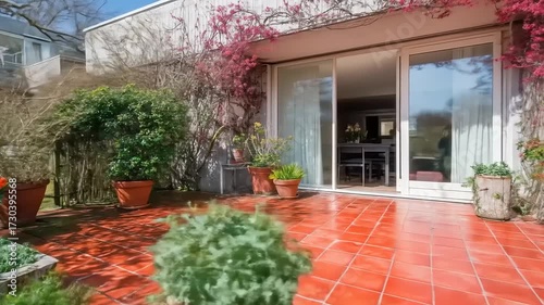 A sunny outdoor patio with a modern home, adorned with vibrant flowers and lush greenery, showcases a beautiful red tile floor.