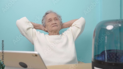 An elderly Asian woman is listening to music from a digital tablet through a wireless speaker.