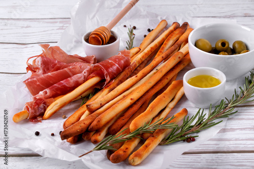 Grissini made of wheat flour with parmesan, rosemary and olive oil. Breadsticks served with olives and prosciutto on white background. Italian crispy snack
