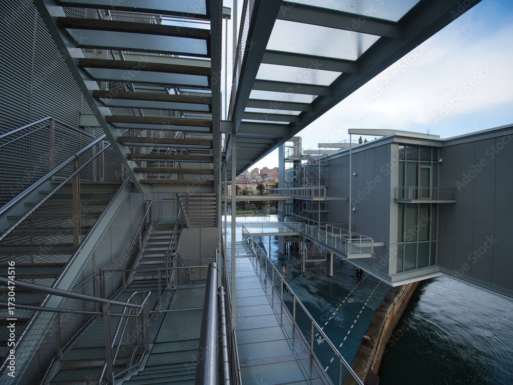 Fototapeta premium Modern Architecture Walkway Over Water - A metal and glass walkway with industrial design leads over water, reflecting the sky and clouds with a futuristic feel.