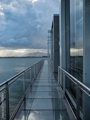 Modern Architecture Walkway Over Water - A metal and glass walkway with industrial design leads over water, reflecting the sky and clouds with a futuristic feel.