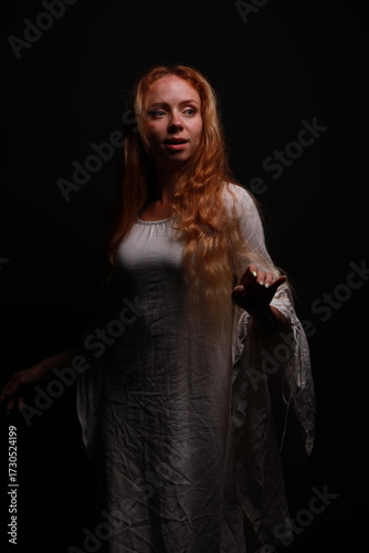 Low key portrait of a young red-haired woman in a vintage white dress in shadows