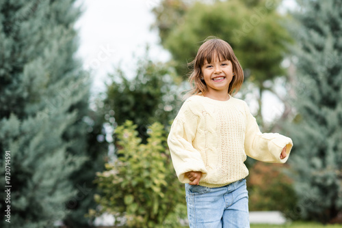 Smiling child girl 4-5 year old wearing knitted sweater and denim pants having fun in park over nature background outdoors. Looking at camera. Autumn season.