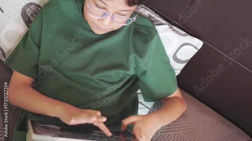 Asian girl focused on playing a game on a digital tablet at home. Top view shot shows childhood engagement with modern technology in her bed for enjoying her leisure time entertainment.
