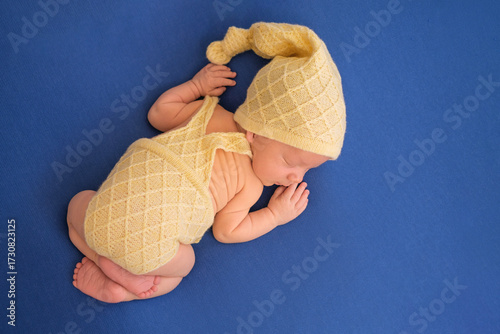 A newborn baby in a knitted yellow suit sleeps sweetly on a blue background. Newborn photo shoot