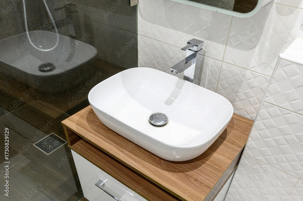 Fototapeta premium A stylish bathroom features a white oval vessel sink atop a wooden cabinet, paired with a chrome faucet. White textured tiles adorn the wall. Reflected shower visible
