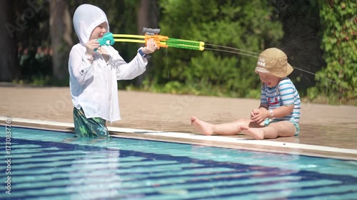 A happy little boy is enjoying himself in the pool. He is playing with a water gun and shooting at his younger brother.