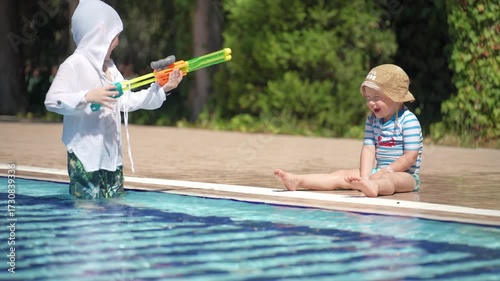 A happy little boy is enjoying himself in the pool. He is playing with a water gun and shooting at his younger brother.