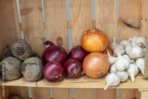 Fresh vegetables including beets, red onions, yellow onions, and garlic arranged on a wooden shelf, showcasing vibrant colors and natural textures in a rustic setting