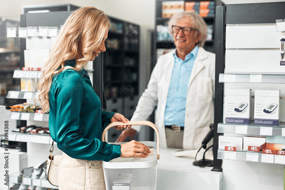 Obraz premium Smiling senior male pharmacist serving customer at modern pharmacy counter