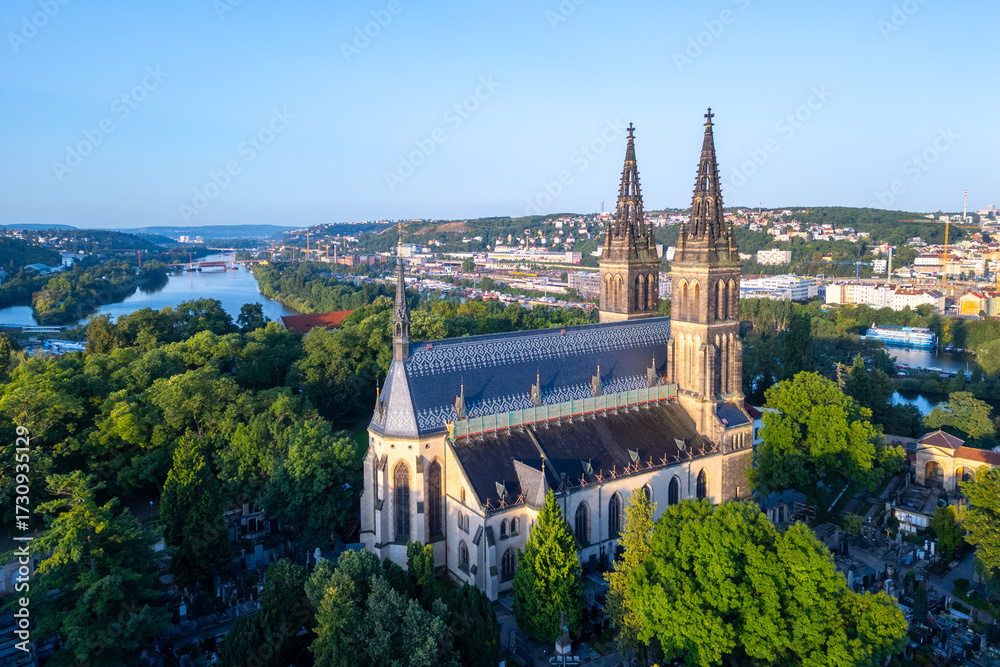 Fototapeta premium Golden sunlight bathes the Basilica of St. Peter and St. Paul in Prague during a tranquil morning. Surrounded by lush greenery, the historic structure stands majestically against the skyline.
