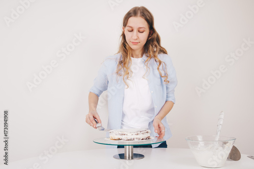 A beautiful young woman with long hair decorating a chocolate sponge cake with a palette knife and butter cream on a special stand to assemble a delicious cake. Home made chocolate sponge cake.