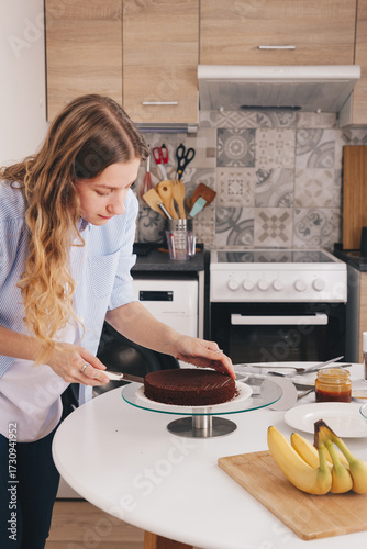A beautiful young woman with long hair cuts a chocolate sponge cake on a special stand to assemble a delicious cake. Home made chocolate sponge cake, traditional cooking, young housewife.