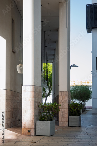 Tall modern columns line a shaded corridor, adorned with elegant hanging lights and framed by neatly trimmed plants, creating a serene and structured architectural walkway.