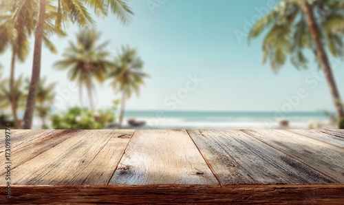 Wooden table sitting on top of a beach next to the ocean, surrounded by lush plants and trees. The sky is filled with white, fluffy clouds, creating a peaceful atmosphere.
