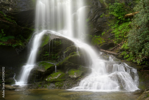 A stunning waterfall flows gracefully over smooth, moss-covered rocks surrounded by lush greenery. Soft morning light enhances the tranquil atmosphere, creating a peaceful scene. Catawba Falls.