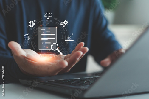 Businessman hand with laptop holding digital document icon with magnifying glass and law symbols. Concept of online contract, document management, compliance and legal technology solutions.