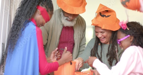 Diverse family filling Halloween buckets at entryway after superhero girl reaching into candy bowl