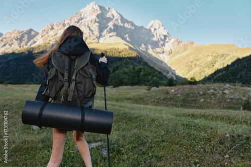 Foto A female hiker walks through a grassy meadow toward rugged mountains, carrying a rolled sleeping mat and a backpack, bathed in clear outdoor light