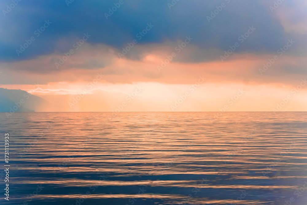 Fototapeta premium Foto pemandangan laut saat matahari terbenam dengan suasana hangat dan tenang. Cahaya matahari oranye keemasan menembus awan gelap dan memantul di atas permukaan air yang beriak lembut, menciptakan nu