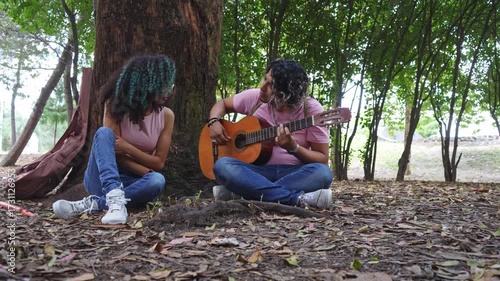 hispanic couple playing guitar sitting in a park, romantic young lovers dating together relaxing with music, boyfriend seducing girfriend singing