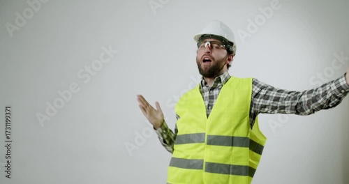 Confused construction worker or foreman shrugging shoulders. He does not know answer. Man with beard wearing hard hat, safety glasses and reflective vest. Isolated studio shot with white background.