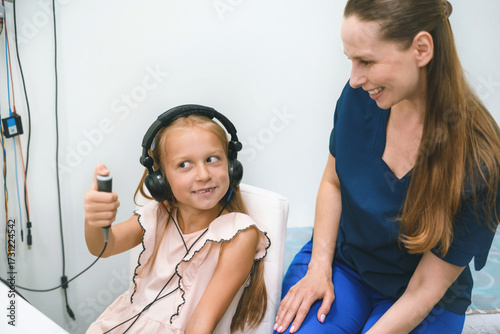 Audiometry hearing test with child and female audiologist doctor at her 40th. Pediatric ear exam in clinic. Hearing healthcare. Hearing loss prevention. Girl patient in headphones clicking button 