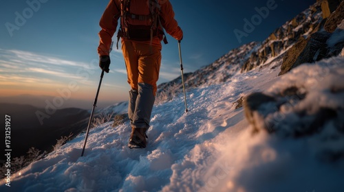 Hiker in orange gear with trekking poles walking uphill on snowy mountain trail at sunrise, adventurous outdoor winter journey in scenic alpine landscape