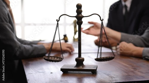Balance scale resting on wooden table with legal professionals discussing contract details during professional meeting inside law office.