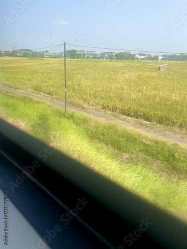 Looking out the window of a moving train. View of rice fields, rural houses and a clear blue sky.