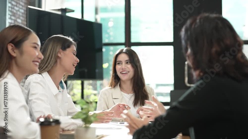 Asian businesswoman in modern office meeting, young female professionals discussing ideas with smiles, brainstorming together in coworking space with natural sunlight and casual business attire.
