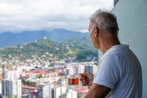 An elderly man drinking tea looks at the green mountain city, standing on the balcony of a skyscraper. The idea of moving to a warm country for retirement