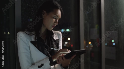 Businesswoman using digital tablet with stylus at night in office, professional Asian female manager working late, illuminated urban background, technology in business, focus and determination.