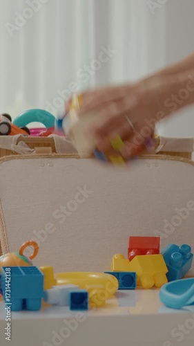 Vertical Screen: A mother gathers various colorful toys scattered around in the living room after her child enjoys a playtime session. The space is bright and inviting, filled with fun items.