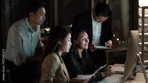 Asian business team working late at night in office, focused professionals using computer and analyzing documents together in dark corporate environment, teamwork productivity in modern workplace