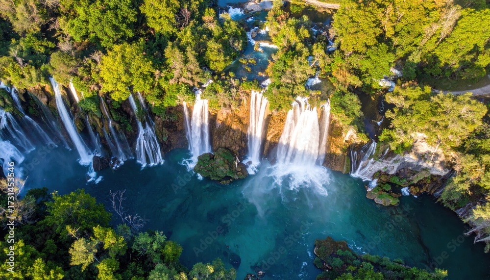 Obraz premium Majestic aerial view showcases a vibrant waterfall cascading down rocky terrain into a turquoise river surrounded by lush green trees, creating a serene and scenic natural environment under bright...