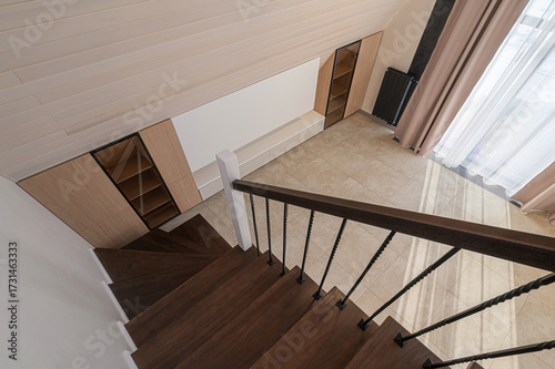 Top-down view of modern loft staircase with dark wooden treads, twisted black iron balusters, light oak slat ceiling, beige marble-effect floor, built-in oak cabinets and shelves, window bench