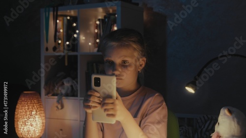 Young girl using smartphone in cozy room at night with soft lighting and decorative items visible
