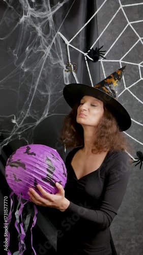 Beautiful witch girl in a witch's cap holds a lilac lantern in her hands against the background of a white web on Halloween