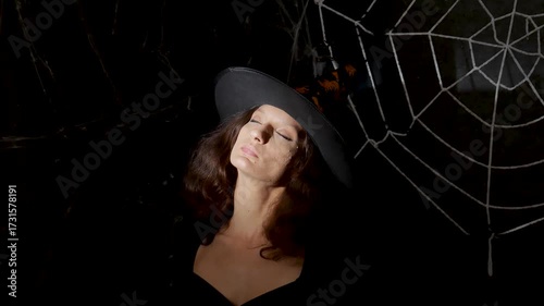 Beautiful girl witch in a witch's cap in the dark under the light closes her eyes against the background of a white web on Halloween