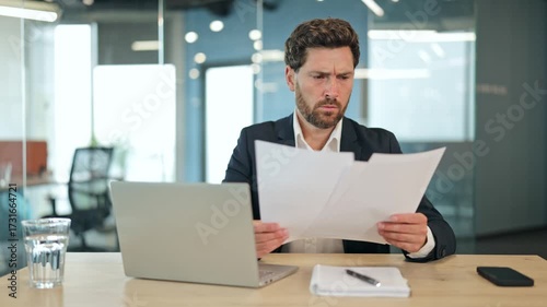 Handsome businessman in a suit analyzes complex documents in a modern office, showing confusion and concern. Diligently reviews papers, processing challenging information on laptop.