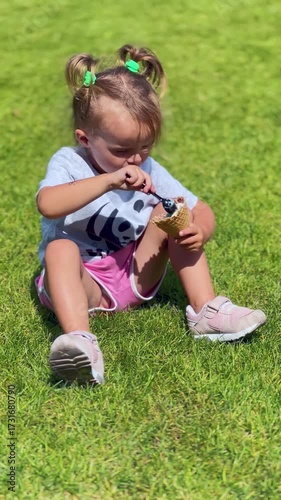 Little girl in pink shorts sits on green grass holding an ice cream cone