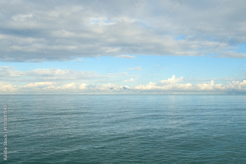 Fototapeta premium Calm ocean reflecting cloudy sky with gentle waves and ripples on water. Sun illuminates clouds in bright blue sky above sea. Beautiful calm magic seascape. Idyllic, picturesque wild nature place.