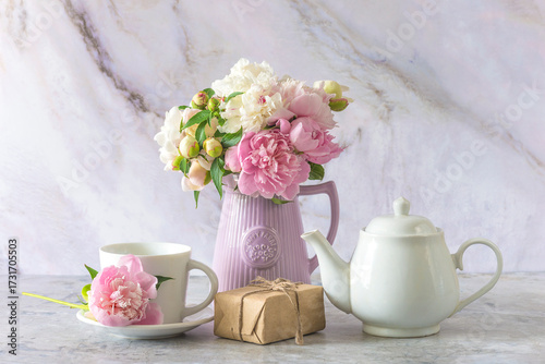 White tea pot, cup of tea, craft gift box and bouquet of pink and white peony flowers on a marbre background