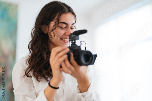 A smiling young woman with dark curly hair holds a professional camera with a microphone, adjusting settings or reviewing footage. She is focused and happy.