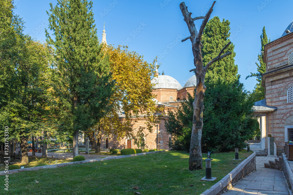Obraz premium Garden view of Muradiye Mosque in Bursa with domes, brick walls, trees and greenery under clear blue sky.