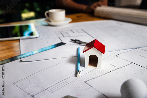 Mini house model on architectural blueprint with pencil, caliper, and tools on wooden desk. Concept of real estate planning, construction, and home design.